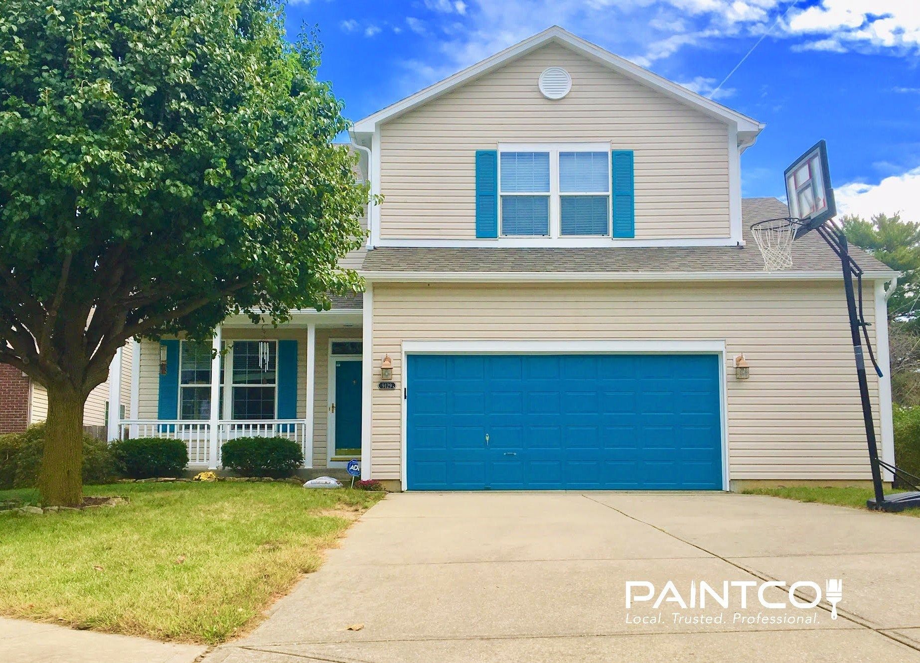 Two-story house with tan siding, teal shutters and garage door, basketball hoop in driveway, and a cloudy blue sky.