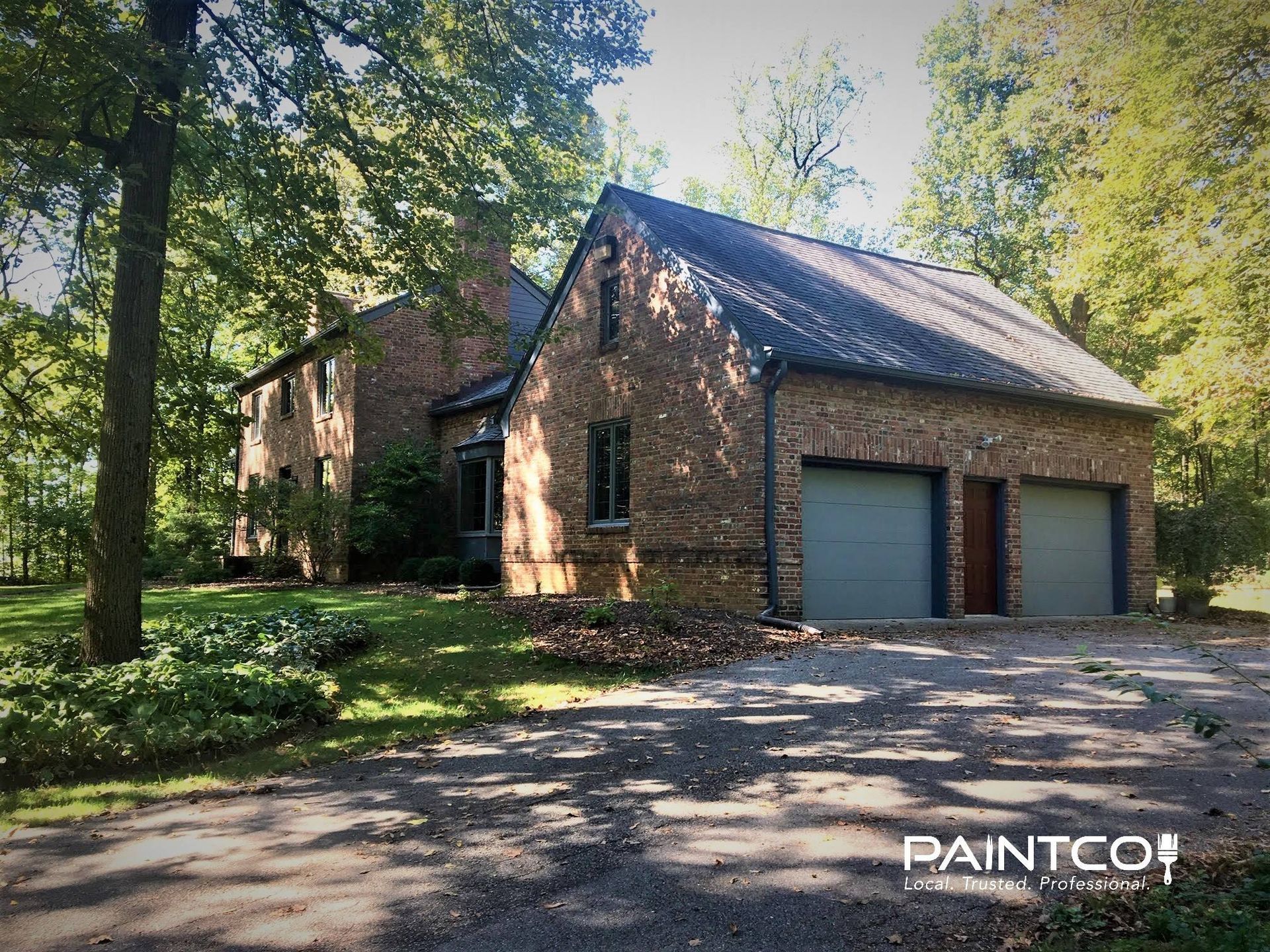 Brick house with two-car garage and green doors; set in wooded area with gravel driveway.