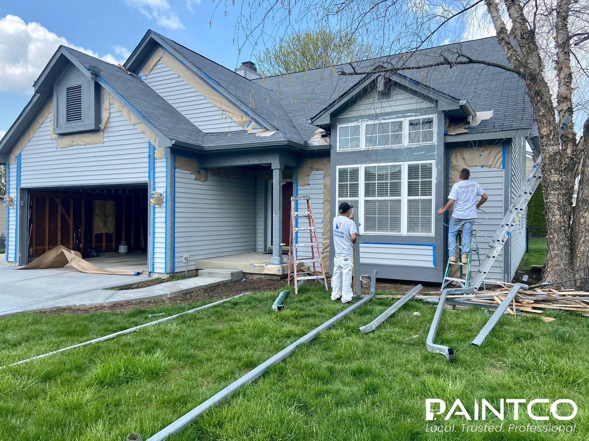 Two painters painting the exterior of a blue-gray house. Ladders and materials are present.