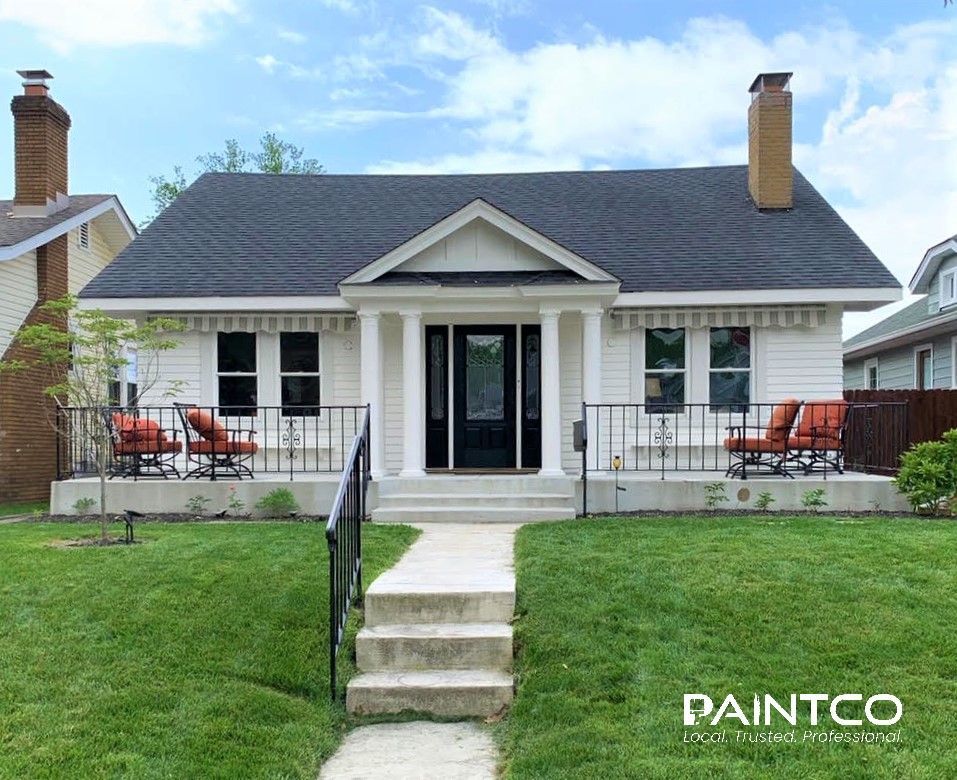 White house with black roof, porch, and small lawn; two chimneys.