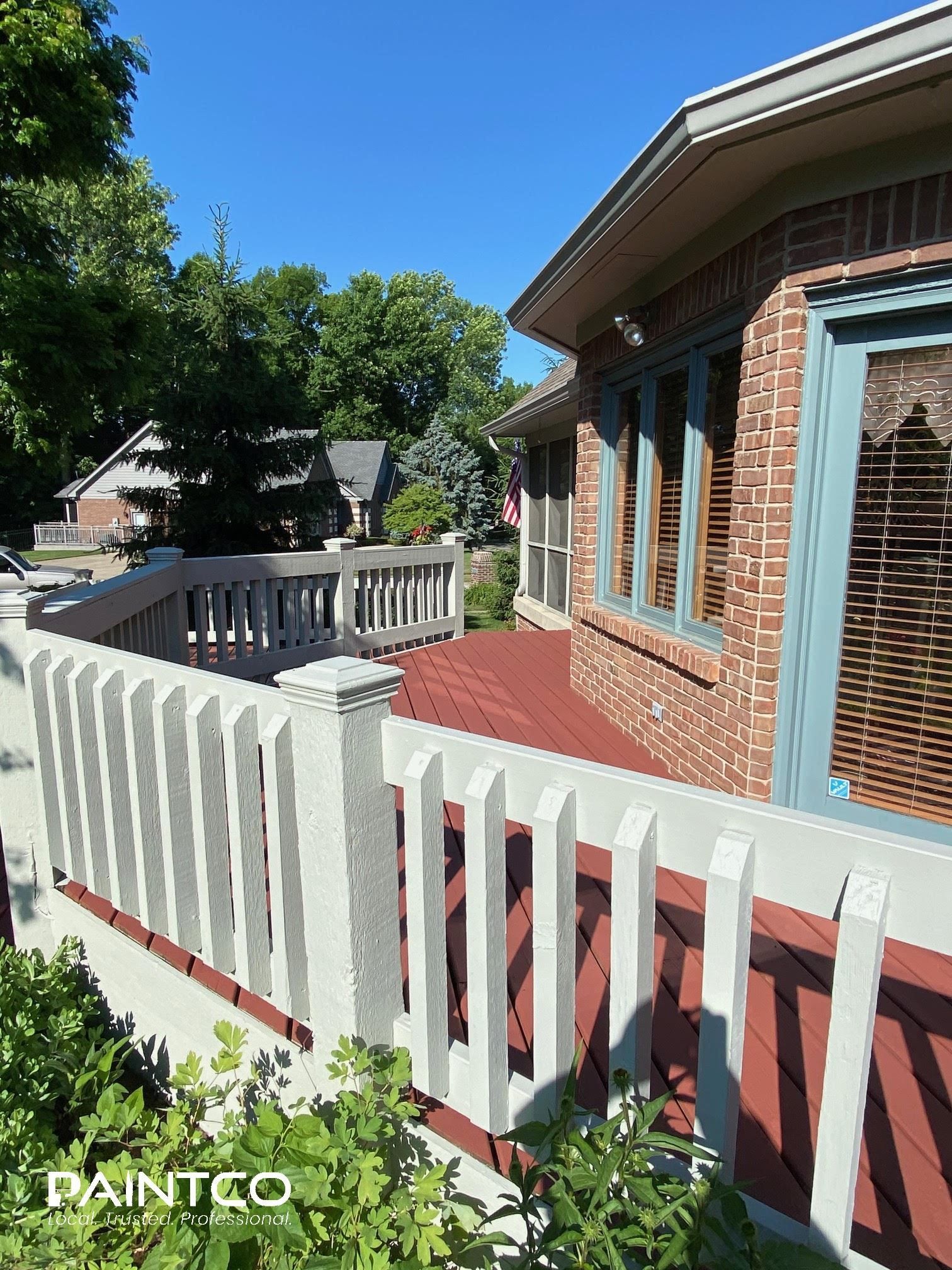 Red deck with white railings, brick house, and blue sky.
