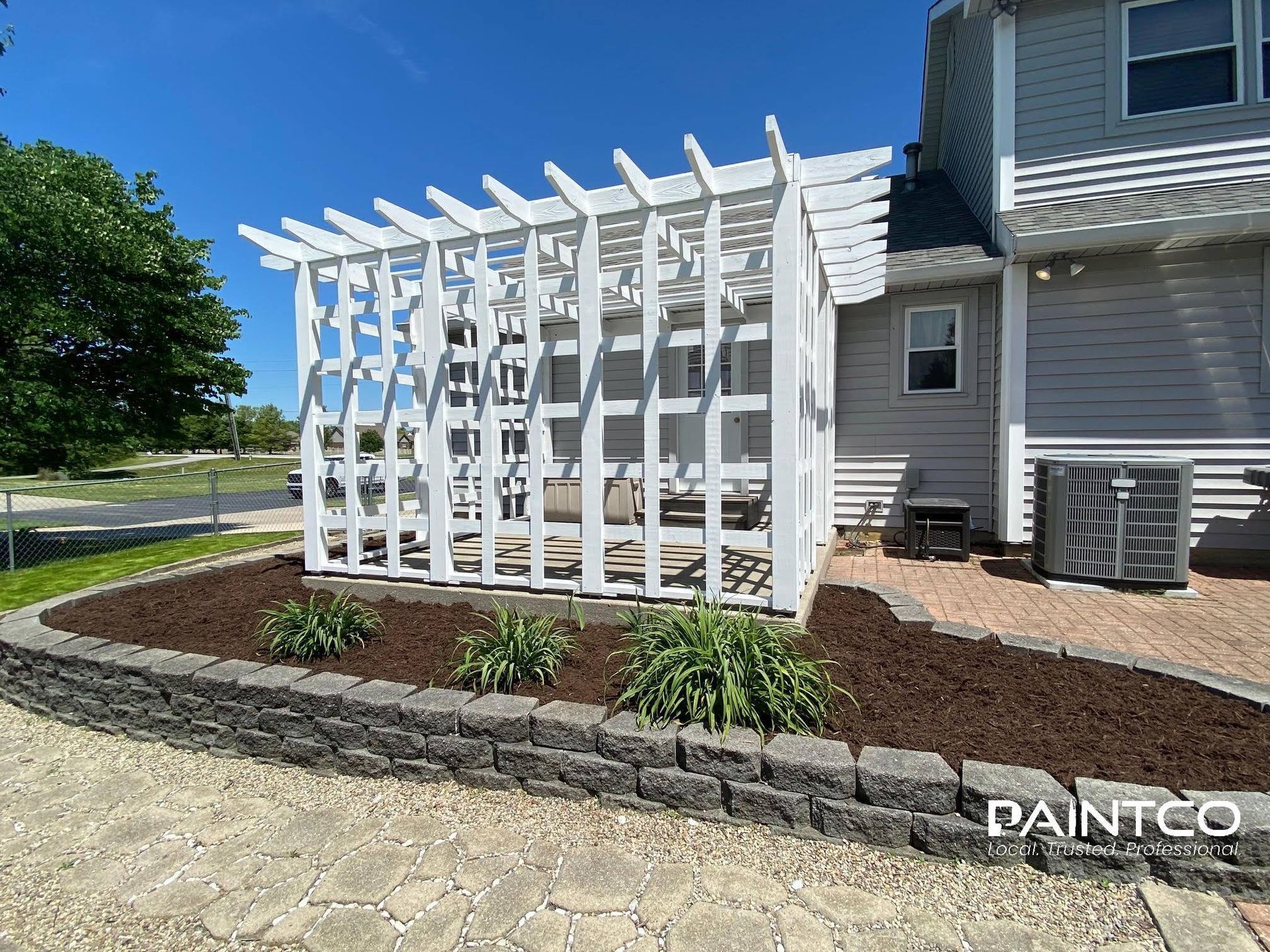 White pergola next to a gray house, with a low stone wall and brown mulch.