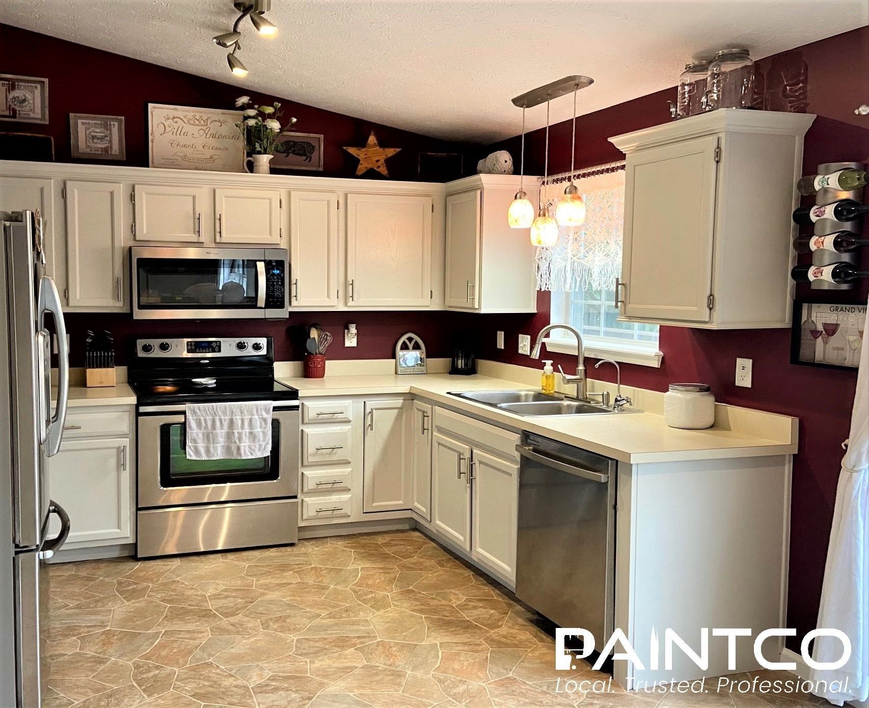 A kitchen with white cabinets, stainless steel appliances, maroon walls, and tan flooring.
