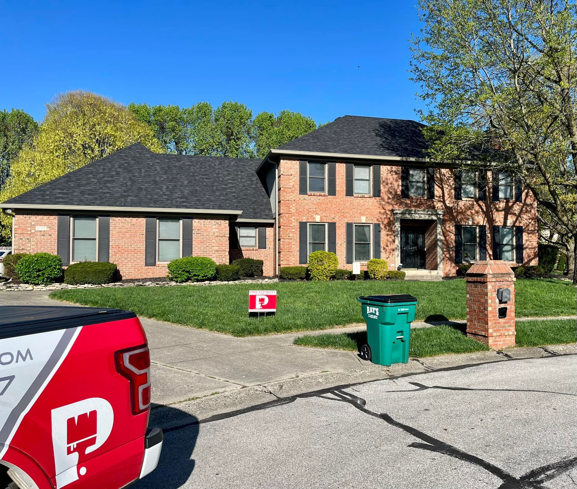 Two-story brick house with black roof, shutters, and lush lawn. A truck with red logo parked in front.