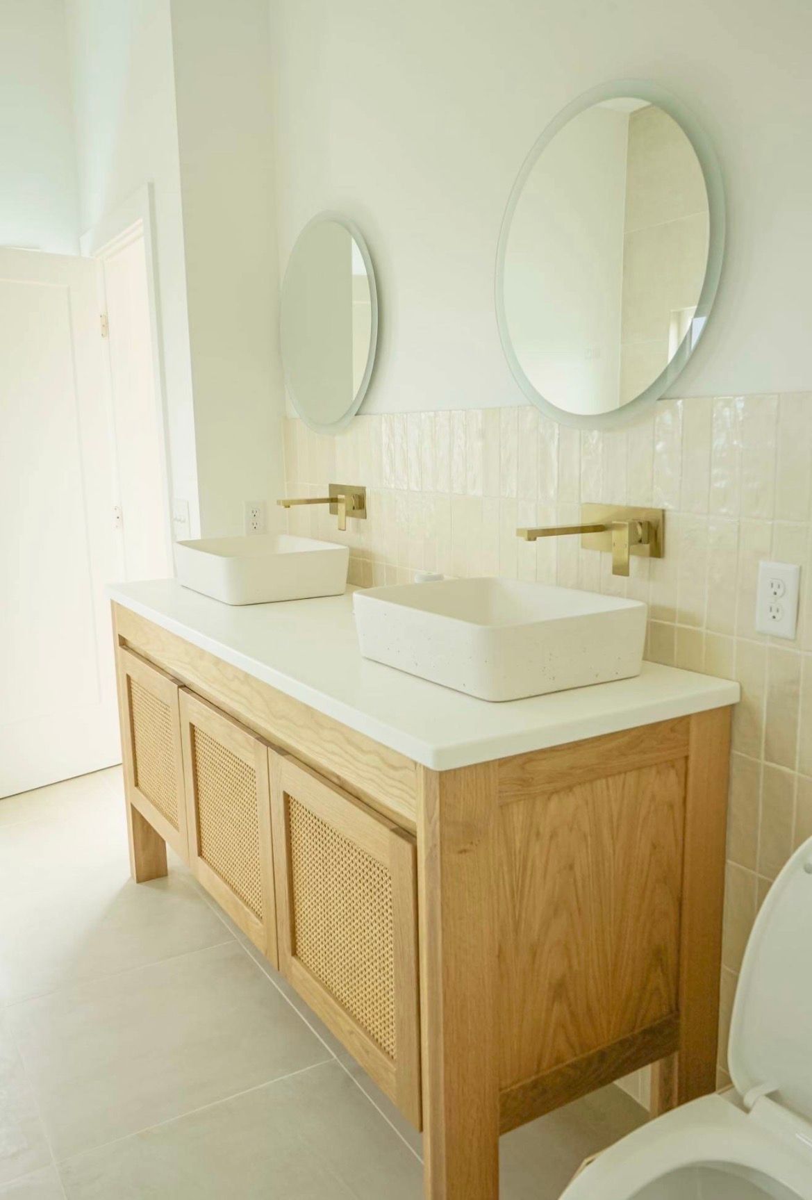 A light wood double vanity with white vessel sinks and gold fixtures, featuring two oval mirrors against tiled walls.