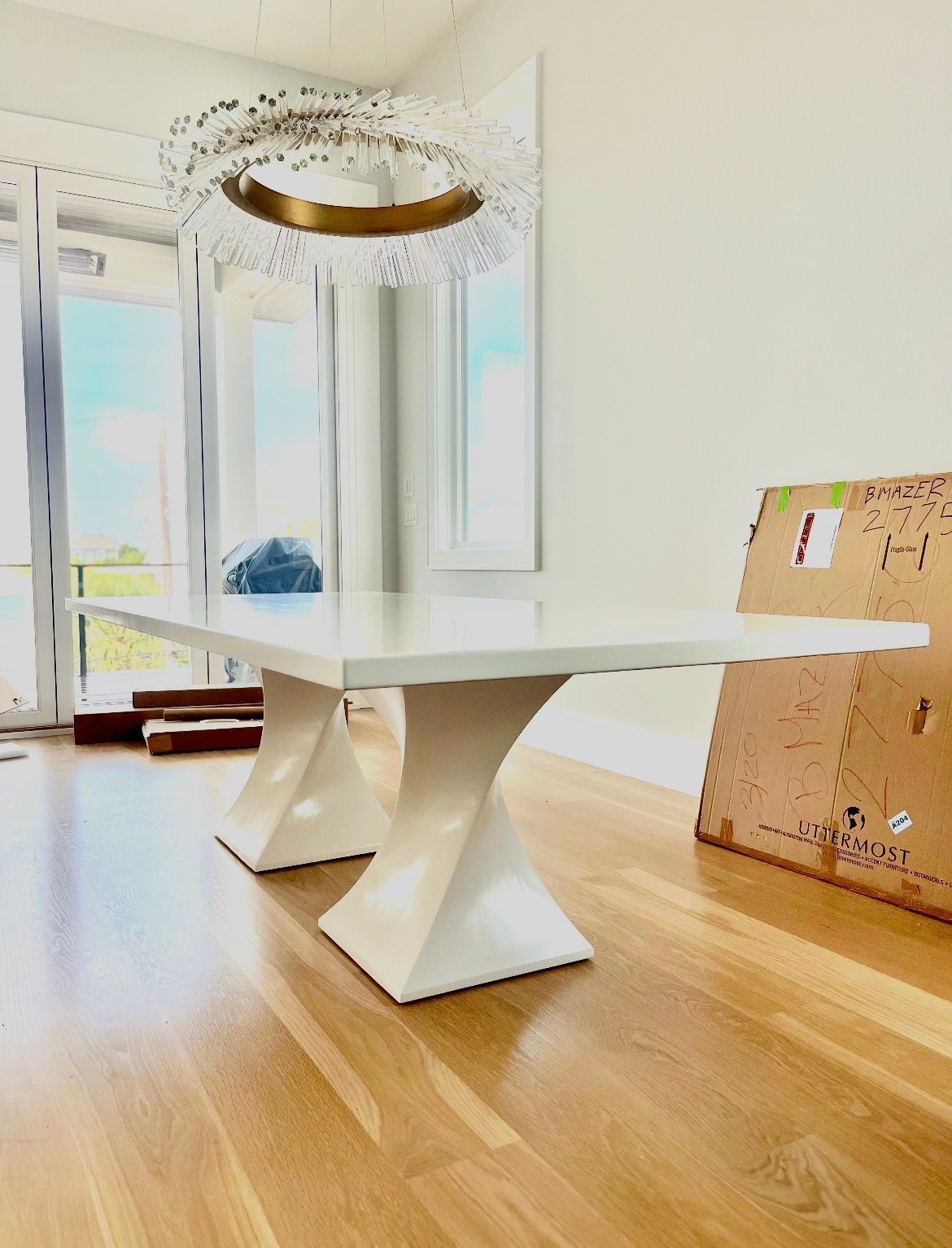 A modern white dining table with two pedestal bases sits on hardwood floors under a circular crystal chandelier.