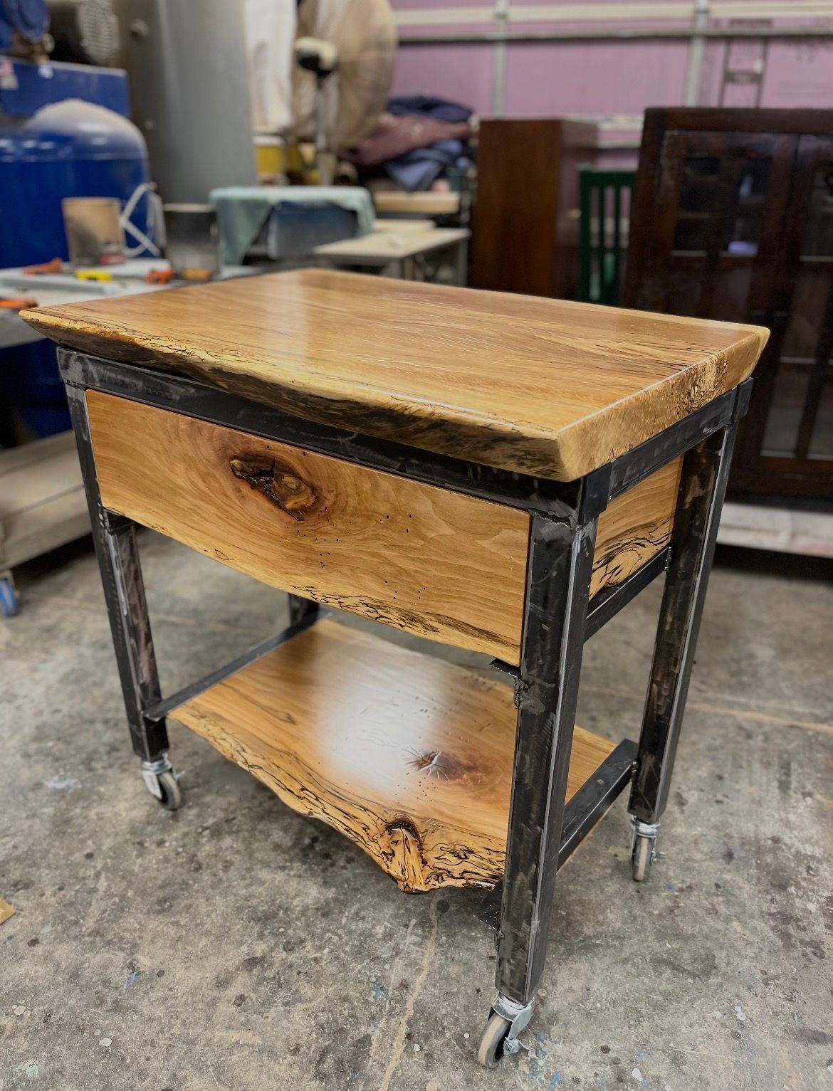 A rolling cart with two natural-edge wood shelves and a single drawer, supported by a rustic dark metal frame.