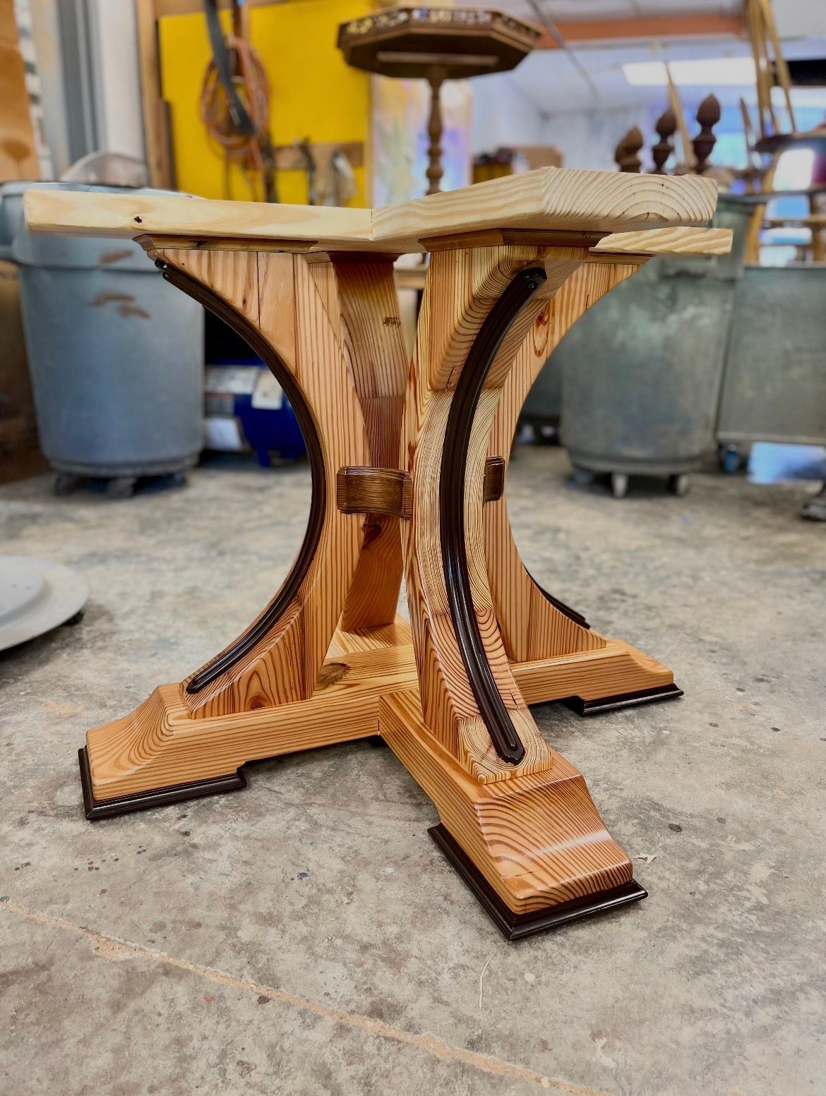 A wooden pedestal table base featuring four curved legs with dark inlaid accents, sitting on a concrete workshop floor.