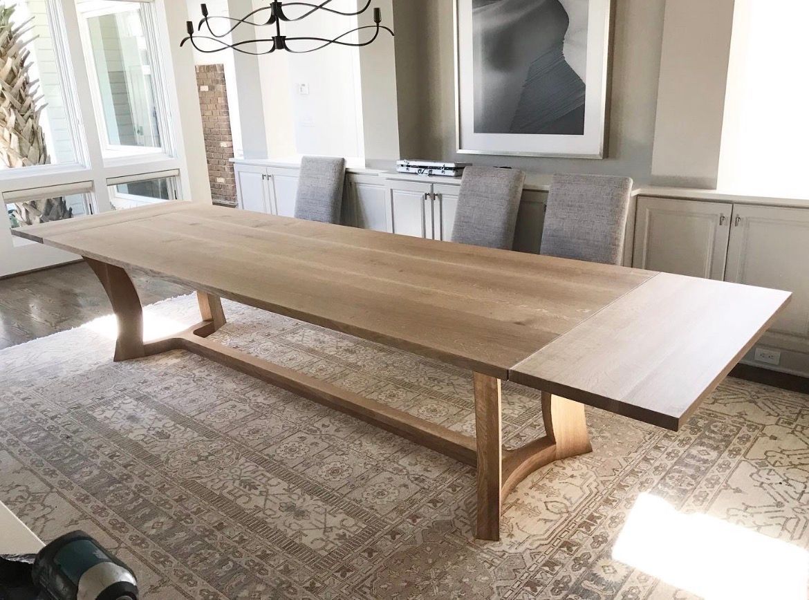 A long, light-wood dining table with curved, trestle-style legs sits on a patterned rug in a bright, modern dining room.