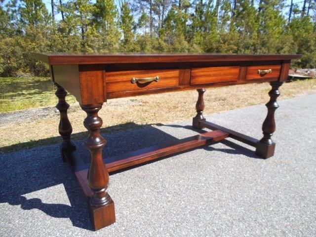 Brown wooden desk with three drawers, turned legs, and a lower stretcher. Outdoors.