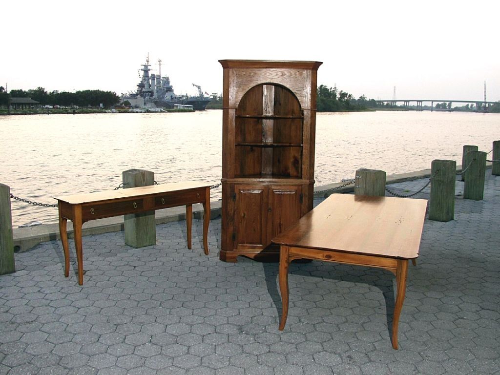 Wooden furniture display outdoors: a corner cabinet, a long table, and a small table on a waterside walkway.