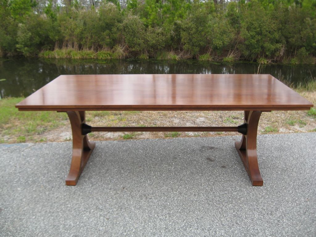 Wooden rectangular dining table on a gravel surface, with a body of water and trees in the background.