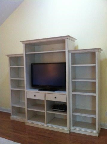 White bookshelf entertainment center with TV in center, shelves on sides, set against a light yellow wall.