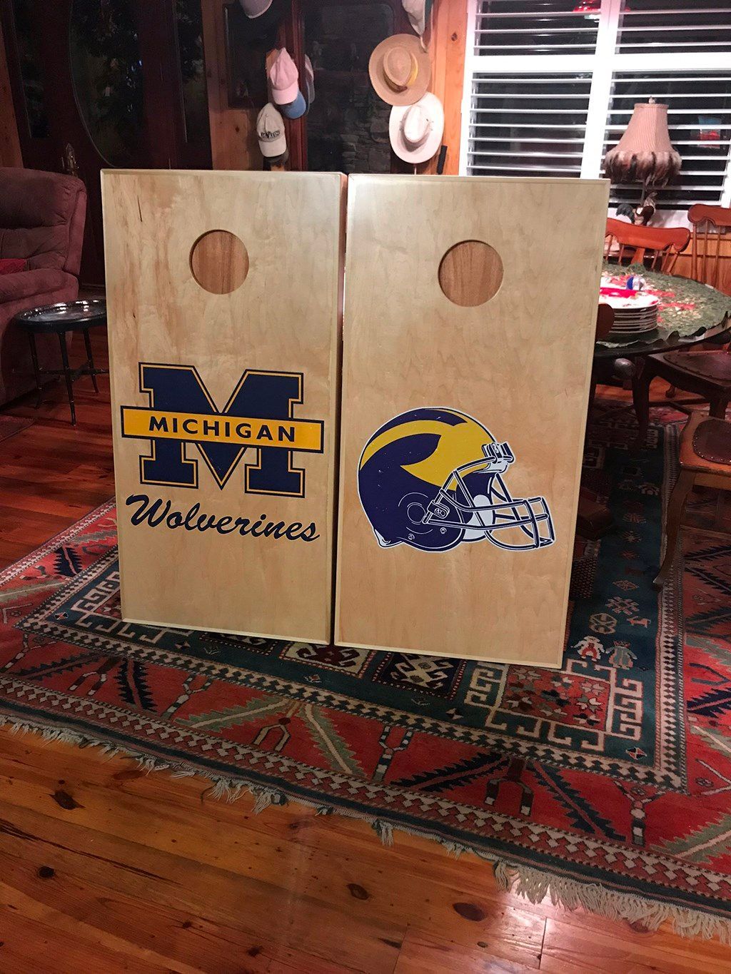 Two Michigan Wolverines cornhole boards on a patterned rug; one with a logo, the other a helmet.