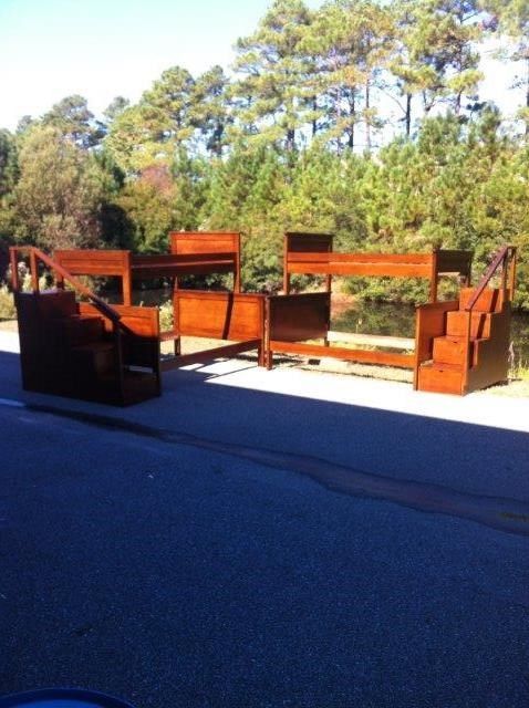 Wooden bunk beds set up on pavement, with staircases and railings. Trees in the background.