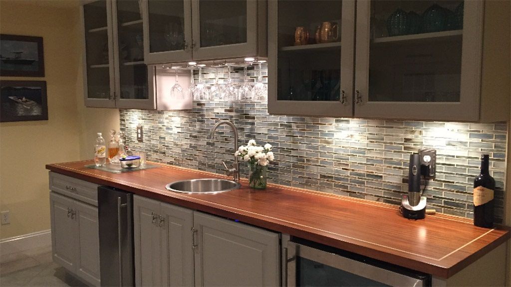 A home bar with gray cabinets, wooden countertop, and a tiled backsplash.