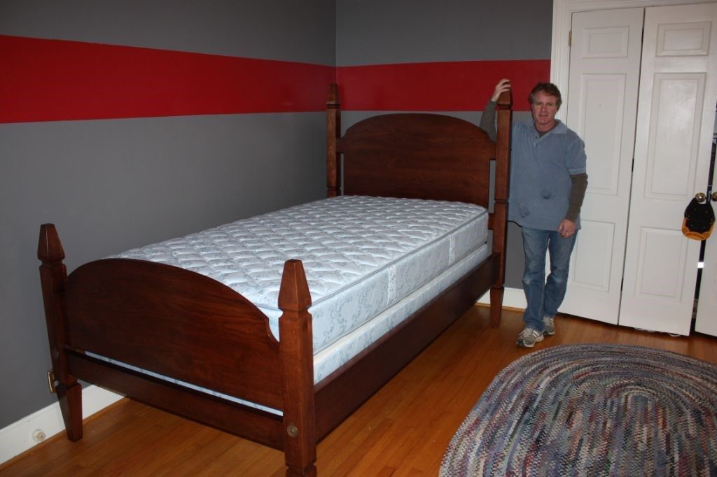 Man stands next to a wooden bed in a room with gray walls and a red stripe.