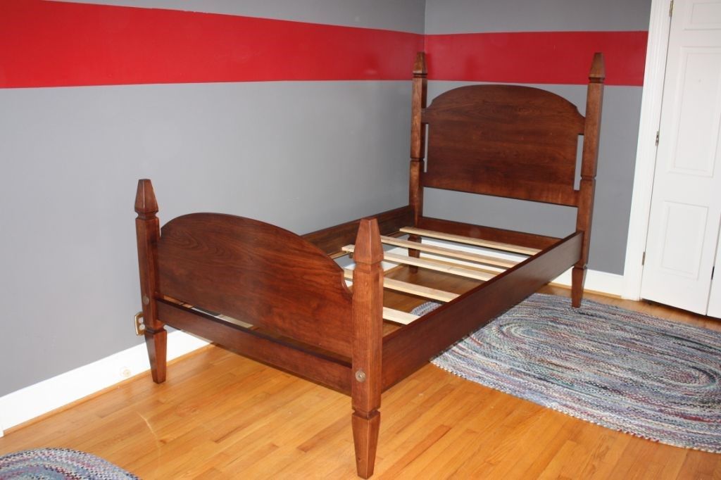 Wooden bed frame in a room with gray and red walls and a braided rug.