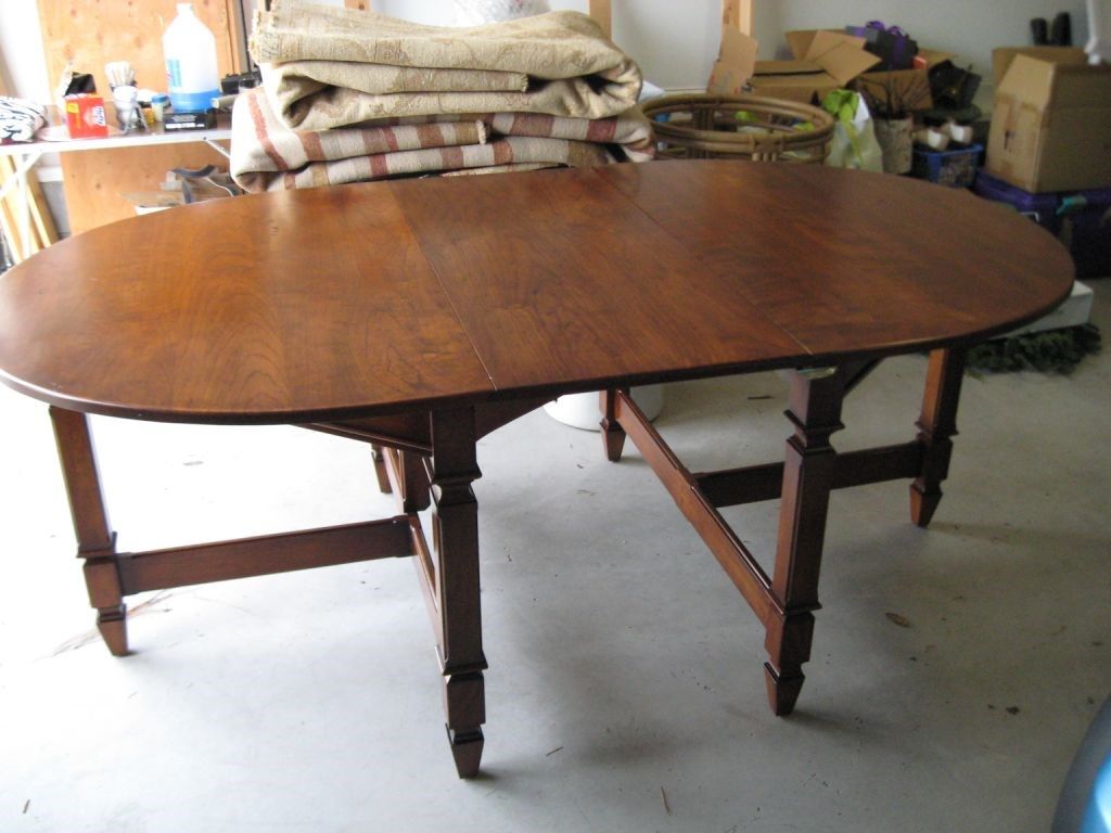 Oval wooden table in a garage, with fold-out leaves, set against a cluttered background.