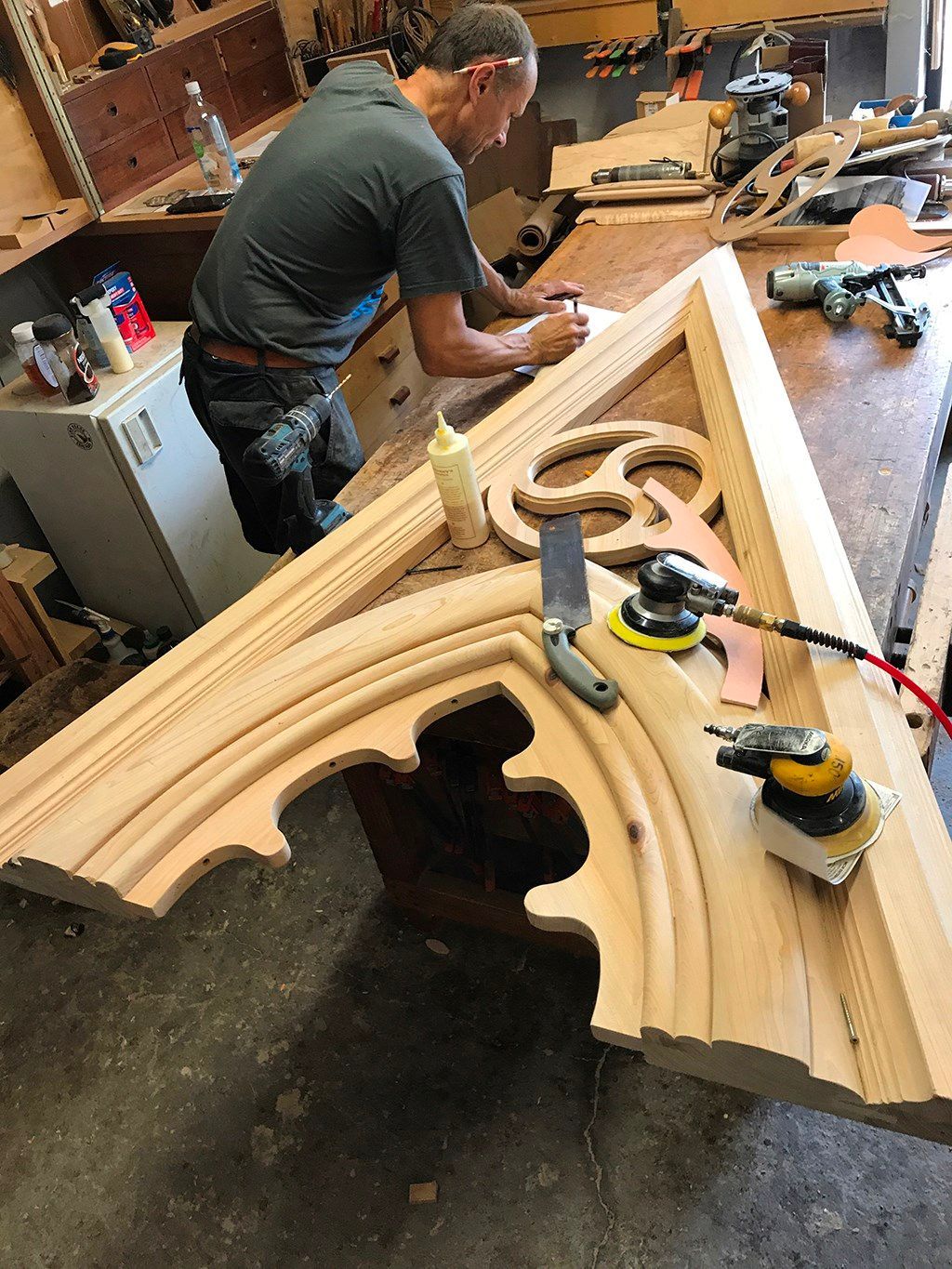 Carpenter working on a carved wooden architectural element in a workshop. Sanding tools and glue visible.