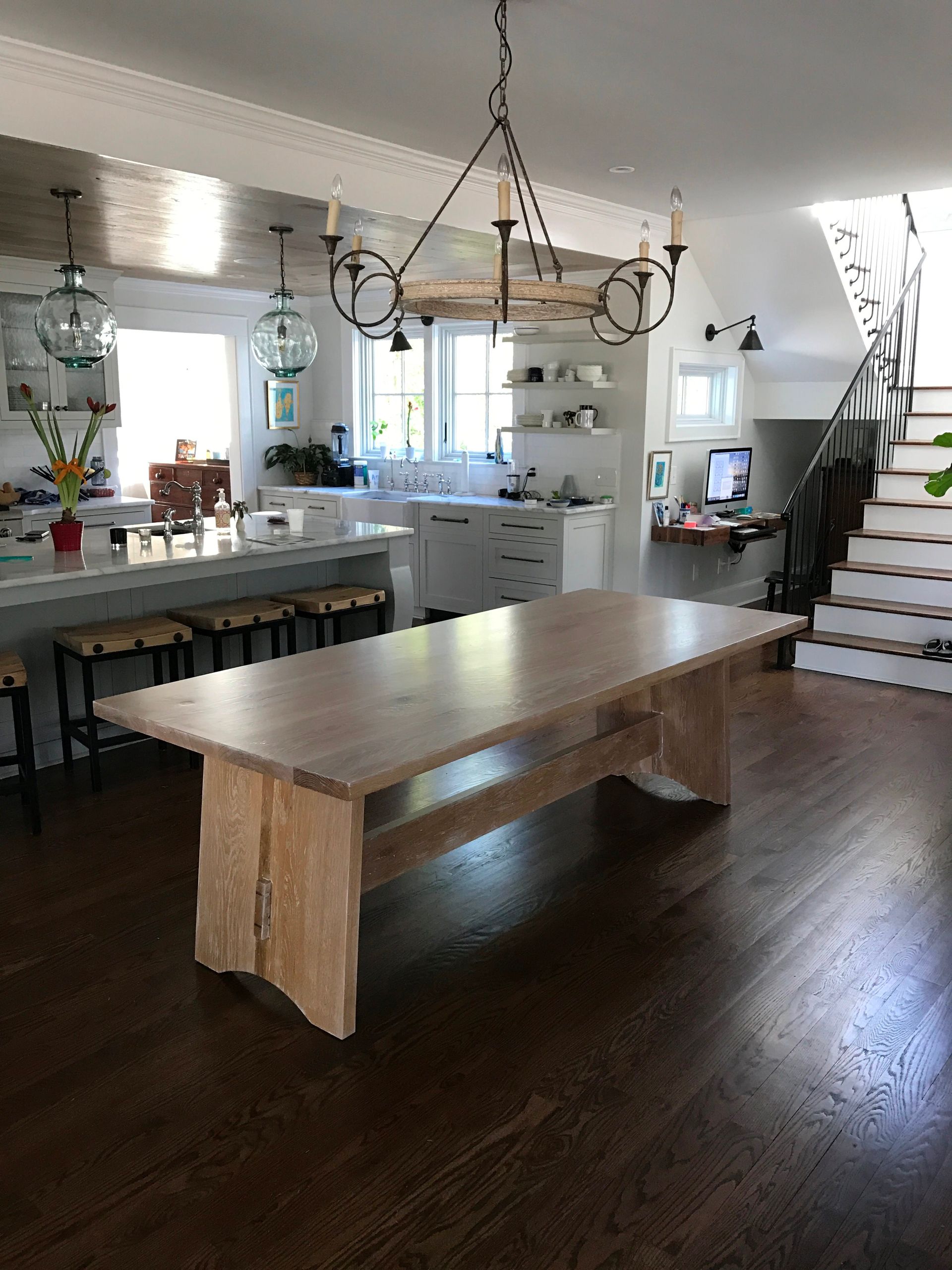 Wooden dining table in a bright room with a kitchen and stairs visible.