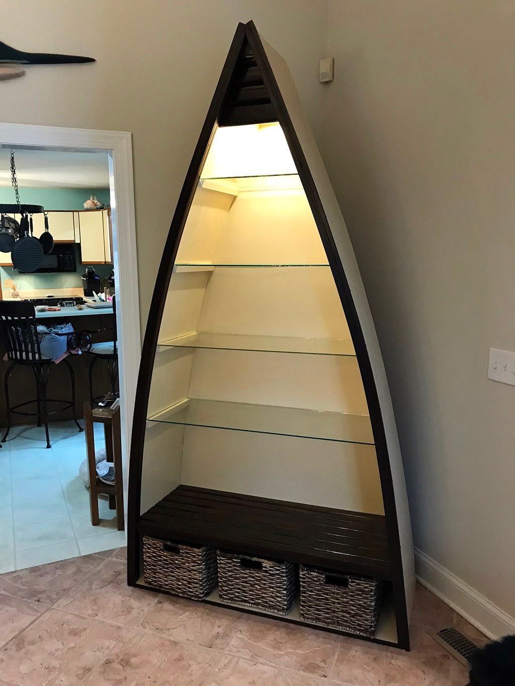 Tall, boat-shaped bookshelf with glass shelves. Dark brown frame, white interior, and three woven storage baskets at the bottom.