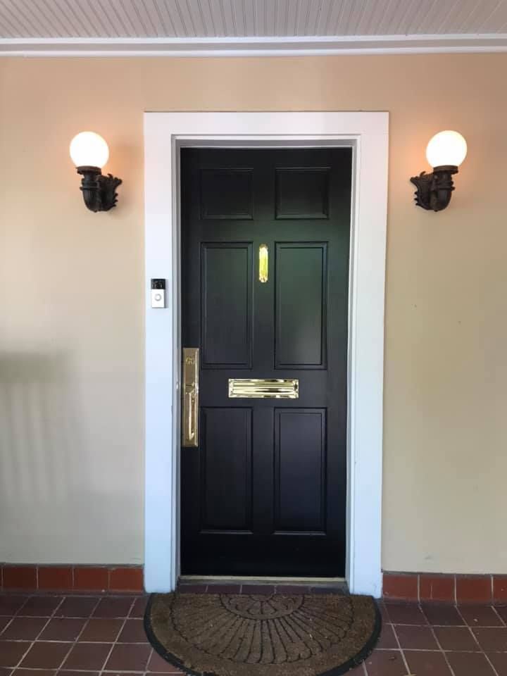 Black front door with white trim, two globe lights, brass hardware, beige walls, and brown doormat.