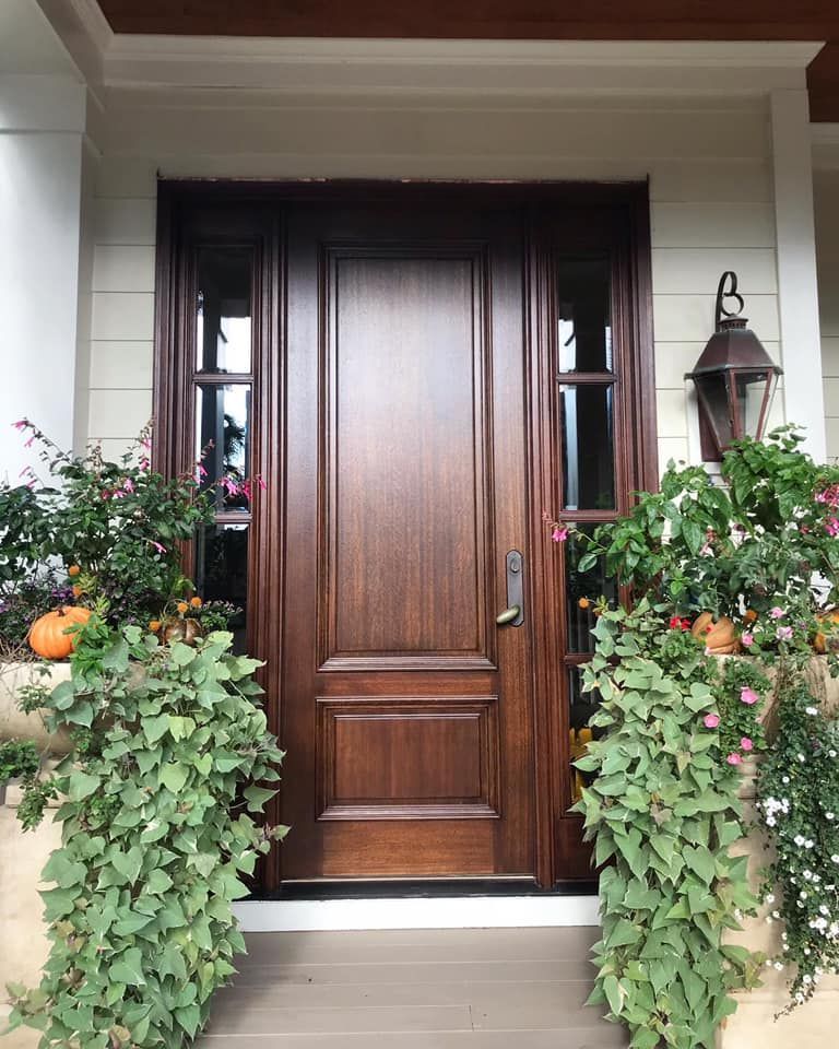 Brown wooden front door with sidelights, flanked by green plants, with a lantern on the right.