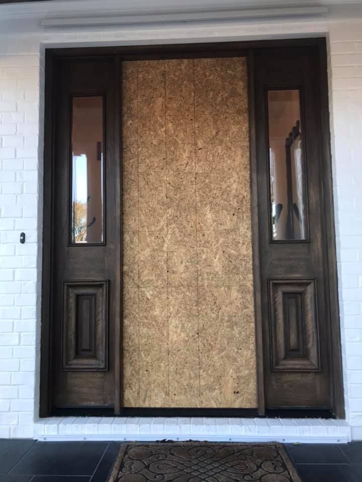 Brown front door boarded up with plywood, flanked by sidelights. White brick exterior.