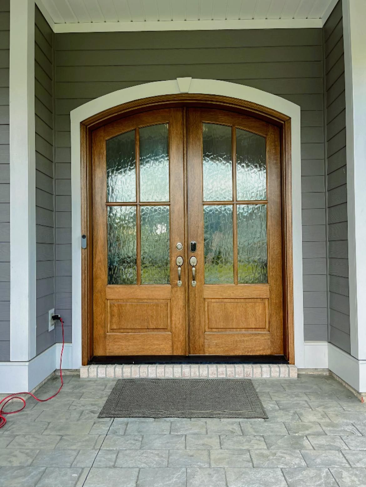 Wooden double doors with arched top and glass panes, set in a gray siding exterior.