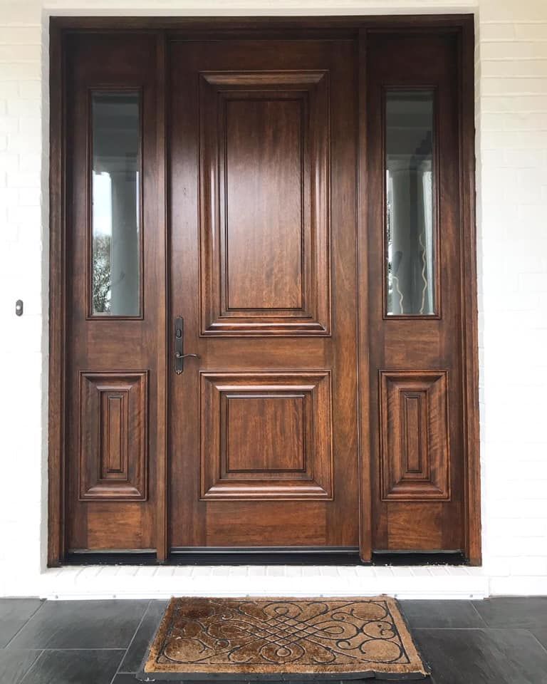 Dark brown wooden front door with sidelights, on a porch with a welcome mat.