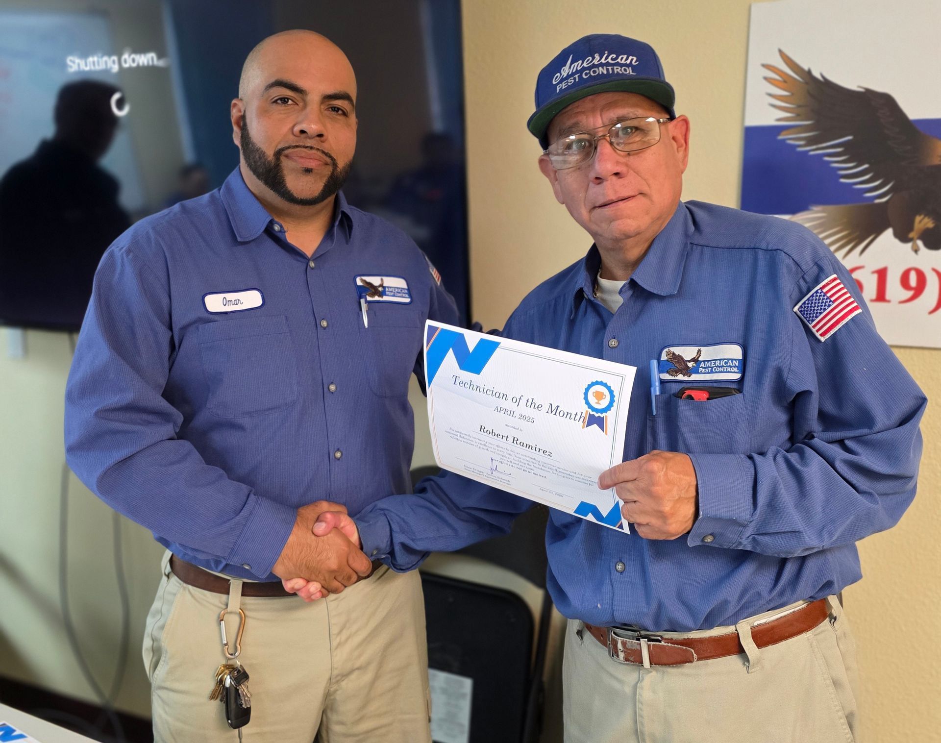Two men in blue shirts shaking hands, one holding a certificate. Background has a wall with an eagle graphic and a shutting down TV.