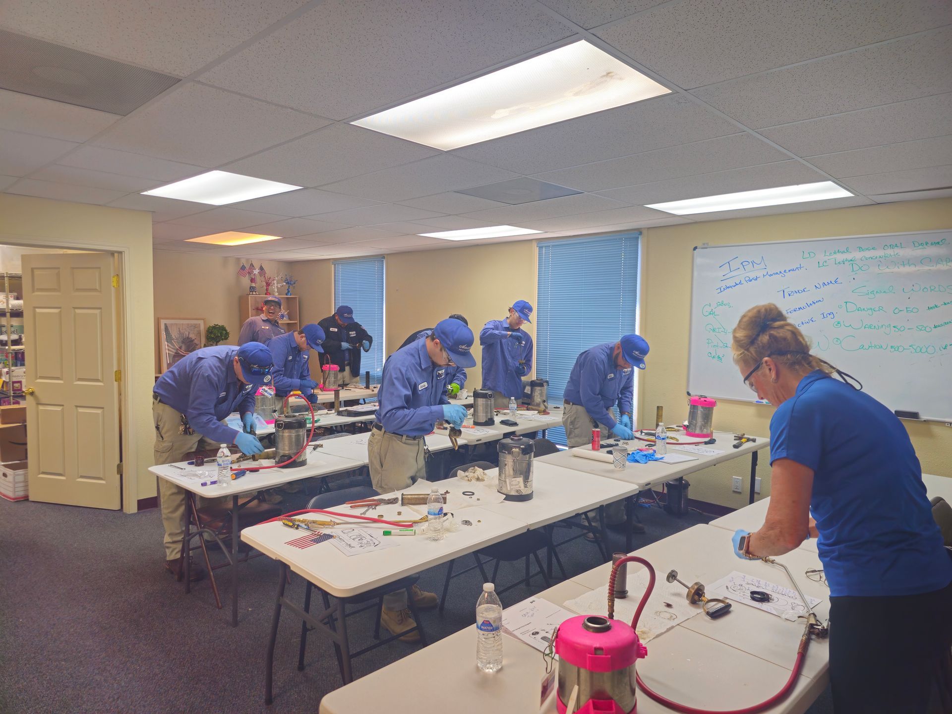 Students in blue uniforms assemble parts at worktables in a classroom. A teacher observes.
