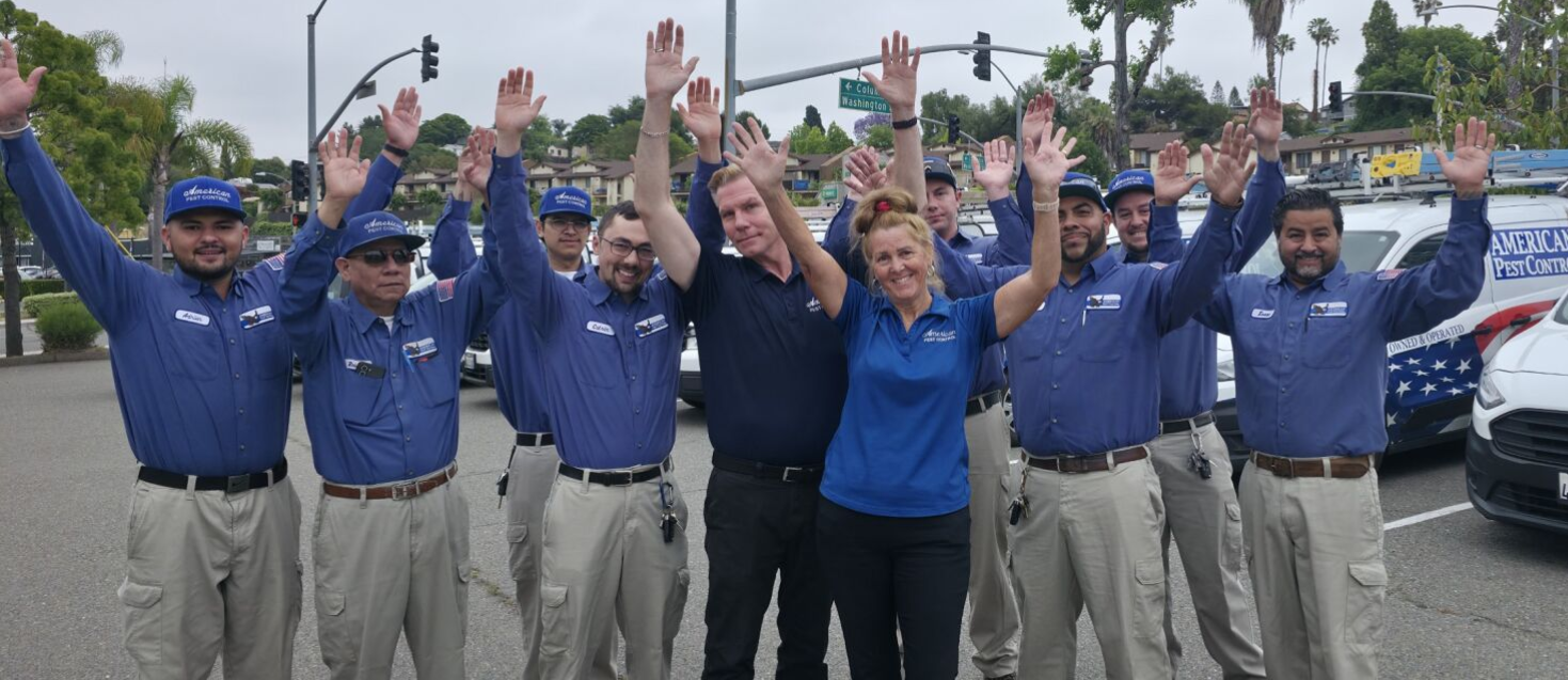 A group of people wearing blue shirts and khaki pants, arms raised, smiling, next to white vans.