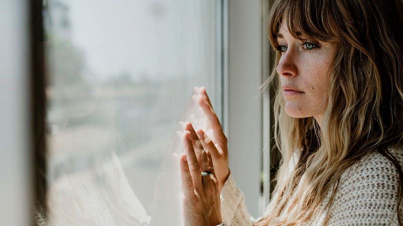 Woman gazing out a window with hands pressed to the glass in a softly lit room