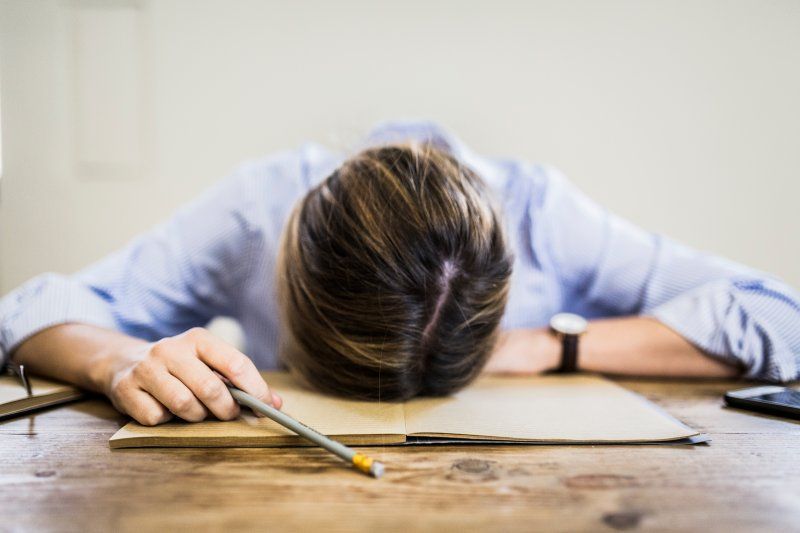 Person slumped face-down at a desk, holding a pencil beside an open notebook.