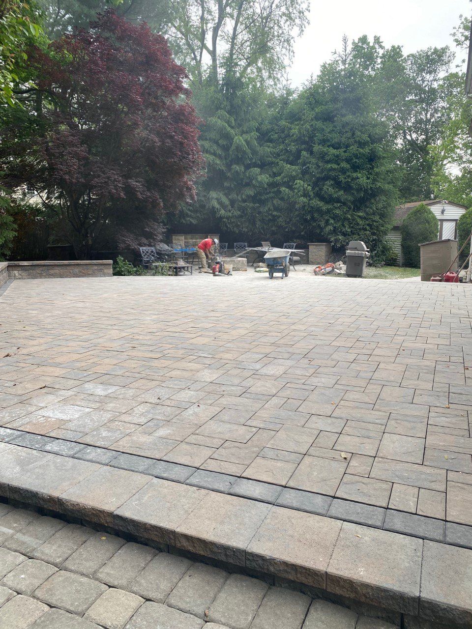 A man is laying bricks on a patio with trees in the background.