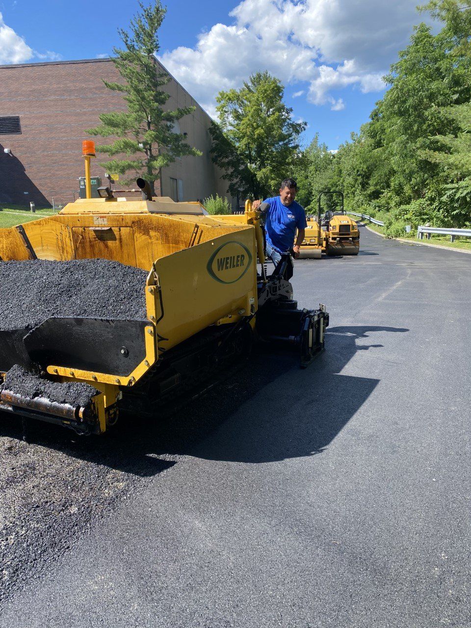 A man in a blue shirt is standing next to a yellow asphalt paving machine