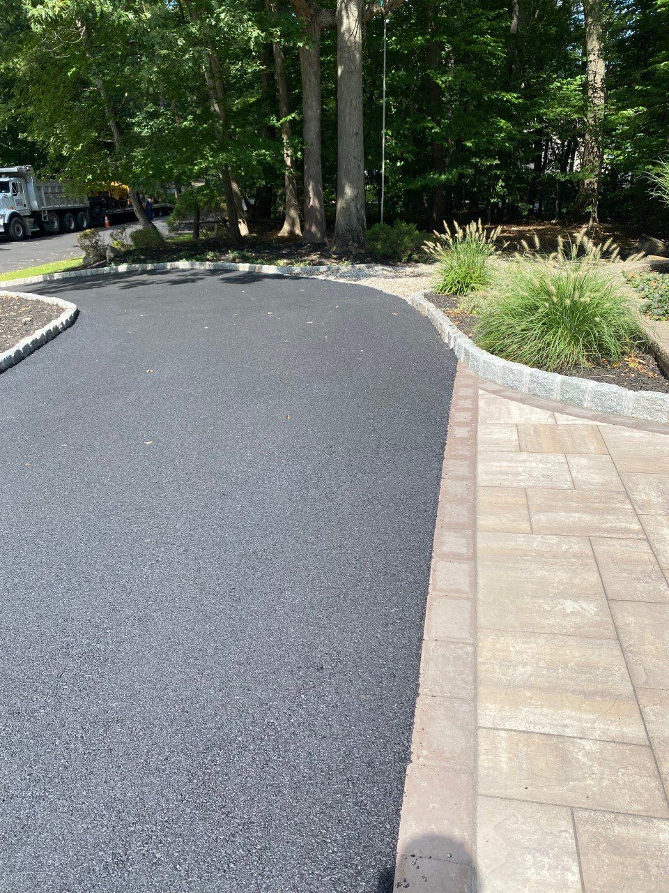 A black asphalt driveway with a brick walkway and trees in the background.