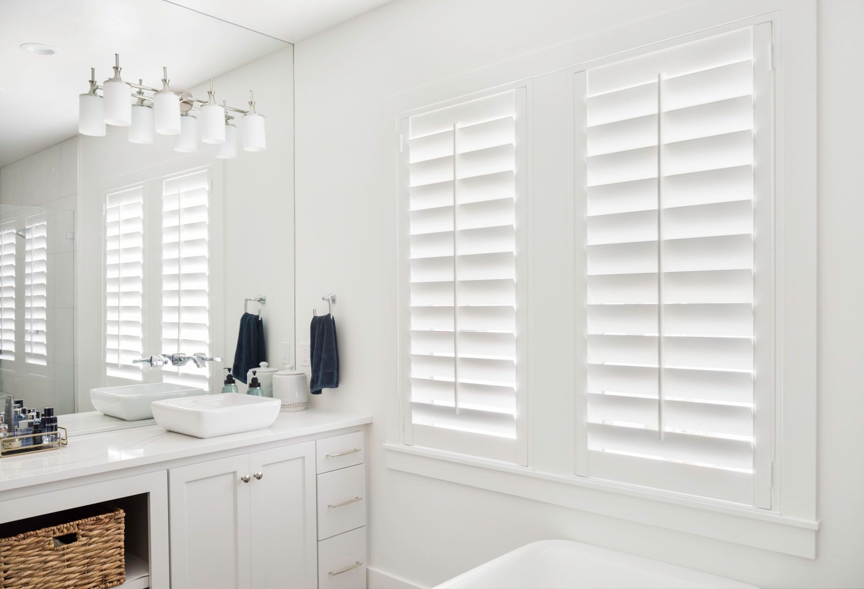 A bathroom with two sinks , a tub , a mirror and white shutters.