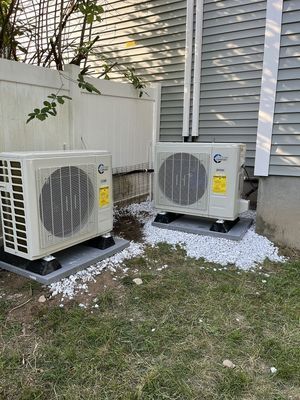 Two beige heat pump units on concrete pads, surrounded by white stones, near a house and fence.