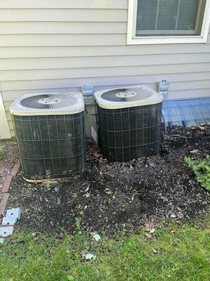 Two dark, outdoor air conditioning units near a building's wall, surrounded by mulch and grass.