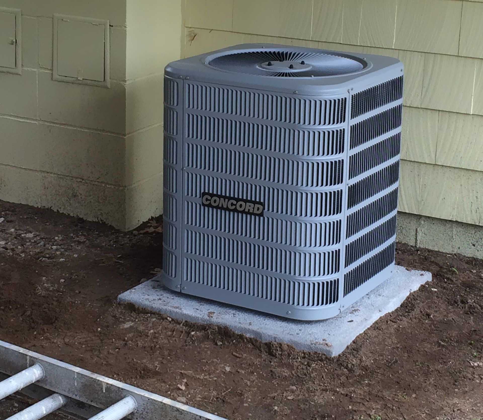 Gray Concord air conditioner on a concrete pad, against a yellow wall.