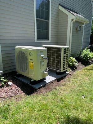 Two beige HVAC units on concrete pads beside a building with gray siding, next to grass.