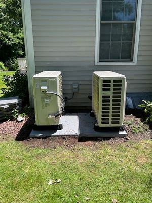 Two beige air conditioning units on concrete pads next to a house with a window; green grass.