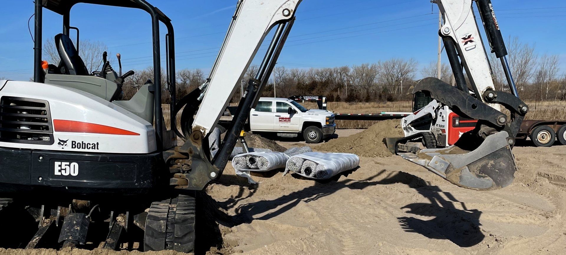 Two excavators lifting a large pipe in a dirt lot, with trucks and bare trees in the background