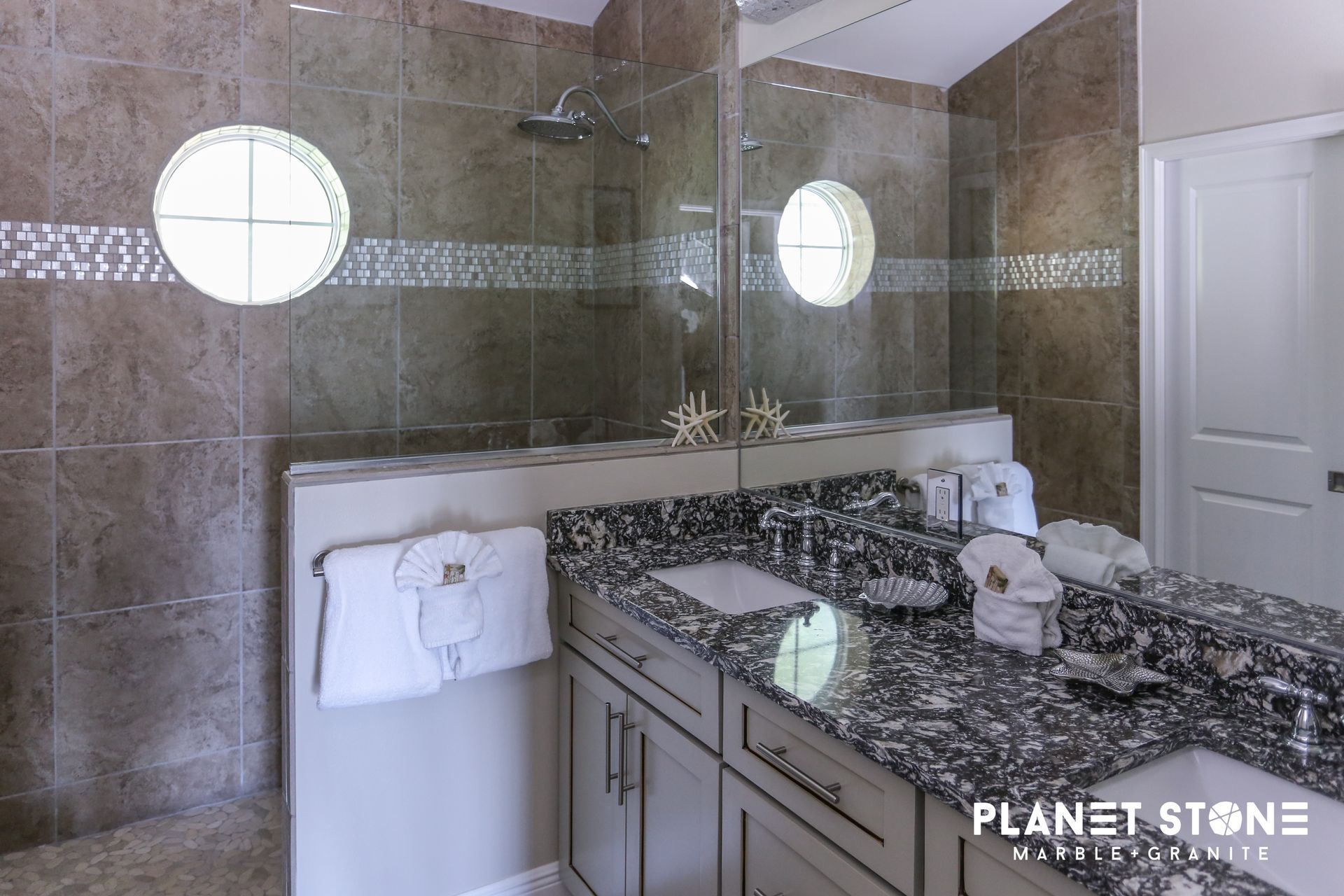 Modern bathroom with granite vanity, large mirror, round windows, and tiled shower walls