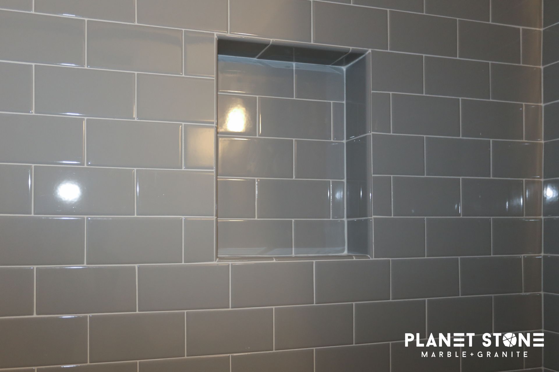 Gray tiled wall with a square mirror and wall sconce, showcasing glossy stone tile sample display