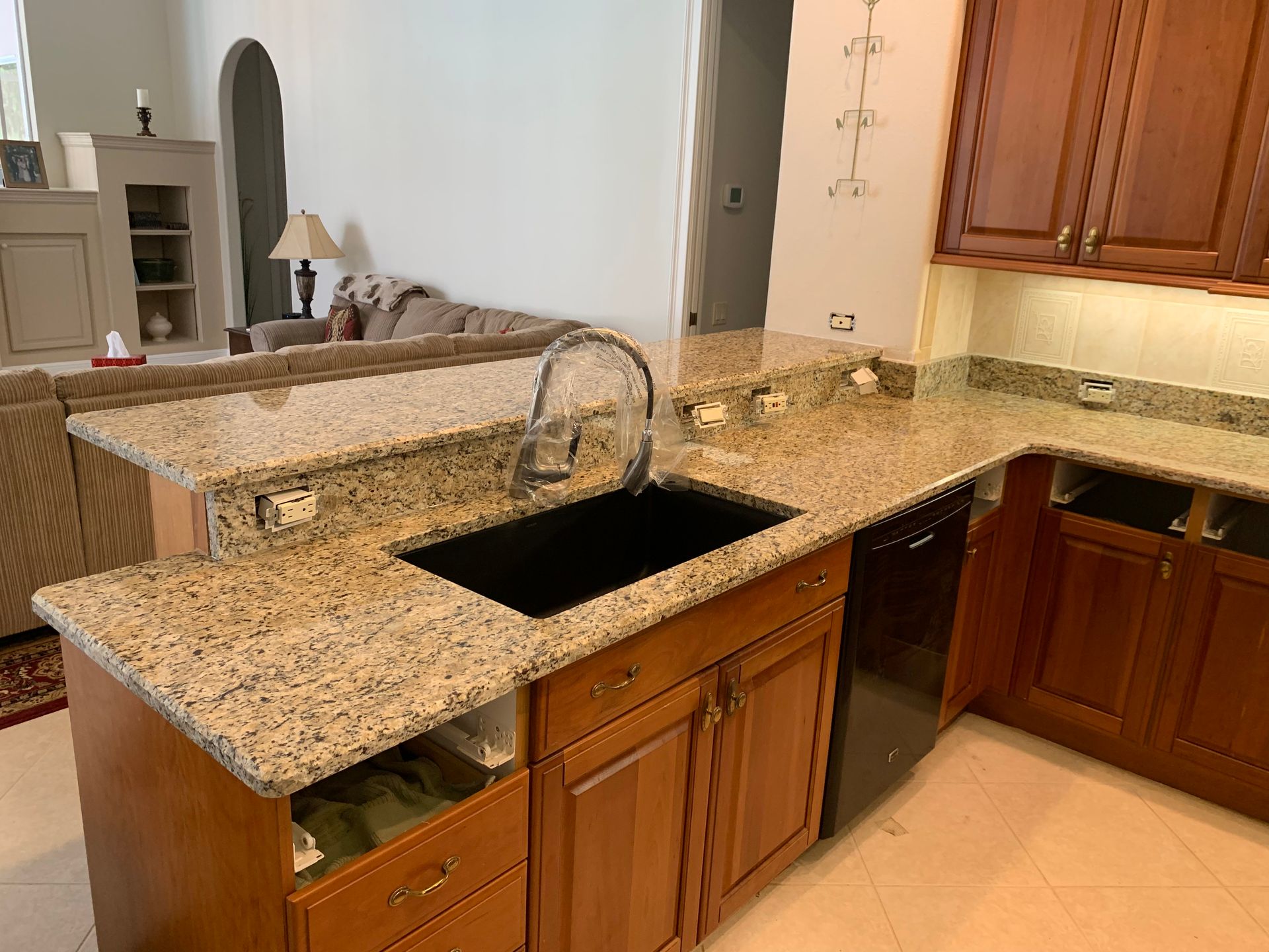 Granite kitchen counter with black sink, wooden cabinets, and breakfast bar overlooking a living room