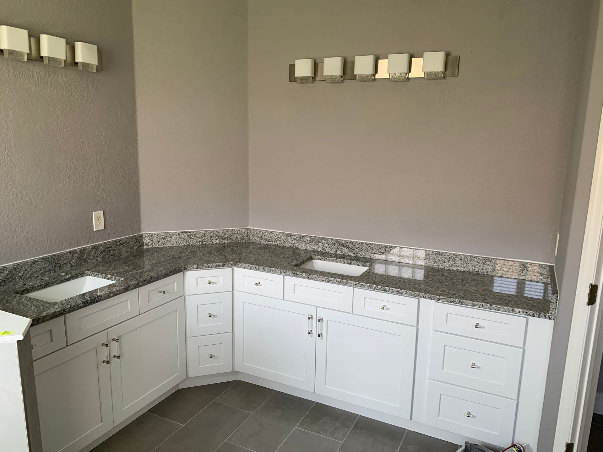 L-shaped bathroom vanity with white cabinets, dark granite countertop, and wall sconces on gray walls